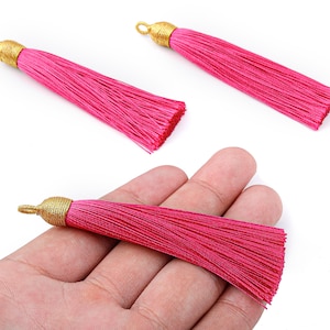 May include: Three hot pink tassels with gold-colored caps and loops. One tassel is held in a hand. The tassels are made of silky threads, perfect for jewelry making or craft projects.