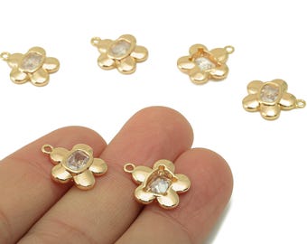 May include: Gold-colored flower-shaped charms with a clear, square-cut center stone. The charms have a loop at the top for attaching to jewelry. Several charms are scattered on a white background, with two held in a hand.