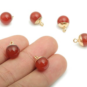 Op de afbeelding: Rode en goudkleurige ronde edelsteenbedels. De bedels hebben een gepolijste, glanzende afwerking. Elke bedel heeft een goudkleurige metalen dop en een kleine lus om aan sieraden te bevestigen. De bedels worden op een witte achtergrond weergegeven.
