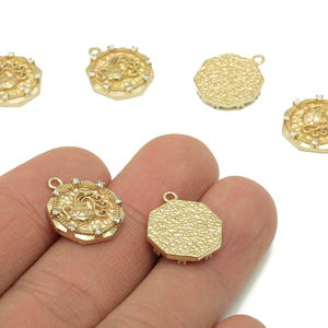 May include: Gold-coloured metal charms with a detailed design. The charms feature a central design surrounded by a decorative border. Some charms have small, clear stones. The charms are octagonal and have a loop for attaching to jewellery.