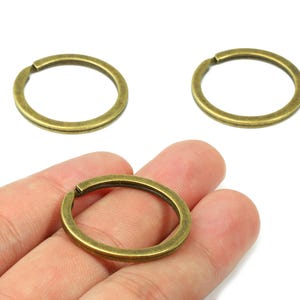 Puede incluir: Tres llaveros de color latón antiguo. Un anillo se sostiene en una mano, mientras que otros dos se colocan sobre una superficie blanca. Los anillos son circulares con una pequeña abertura para sujetar llaves u otros objetos.