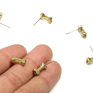 Puede incluir: Pendientes de botón dorados con forma de hueso. Los pendientes son pequeños, con un poste para perforar. Varios pendientes están esparcidos sobre un fondo blanco, algunos sostenidos en una mano.