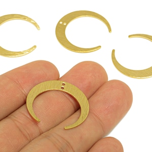 May include: Gold-colored crescent moon charms with a textured surface. The charms have two small holes near the top for stringing. The image shows several charms, with one held in a hand.