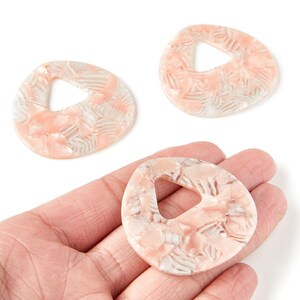 Peut inclure: Trois pendentifs abstraits en forme organique avec une découpe triangulaire centrale. Les pendentifs présentent un motif marbré dans des tons pêche et blanc. Un pendentif est tenu dans une main, mettant en valeur le design et la couleur.