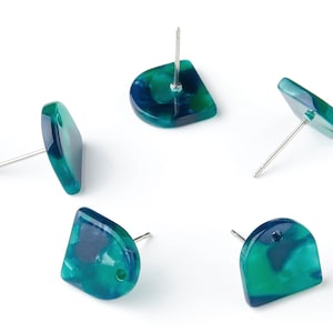 Puede incluir: Cinco pendientes de botón con forma geométrica redondeada. Los pendientes presentan un efecto veteado en tonos turquesa y azul oscuro. Cada pendiente tiene un poste plateado. Se muestran sobre un fondo blanco.