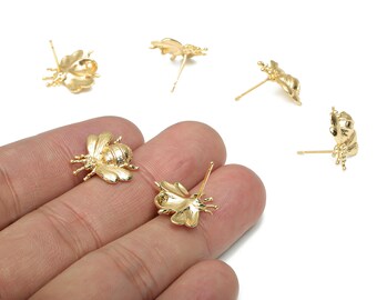 May include: Gold-colored bee-shaped stud earrings. The earrings are detailed with wings, antennae, and legs. Several earrings are scattered on a white background, with two held in a hand. The earrings are made of a shiny metal.