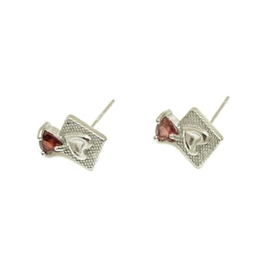 May include: A pair of silver earrings with a small red heart-shaped gemstone and a larger silver heart on a square base.