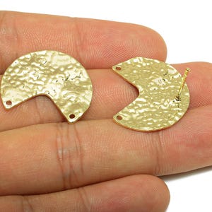 Può includere: Due componenti di orecchini a forma di mezzaluna color oro con una texture martellata. Ogni pezzo ha due piccoli fori alle estremità e un perno sul retro di uno. Gli orecchini sono esposti su una mano.