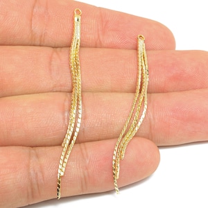 Puede incluir: Dos componentes de pendientes dorados. Cada uno tiene un pequeño lazo en la parte superior y múltiples hebras de delicadas cadenas onduladas. Las cadenas tienen una superficie texturizada y reflectante. Los componentes se sostienen en una mano.