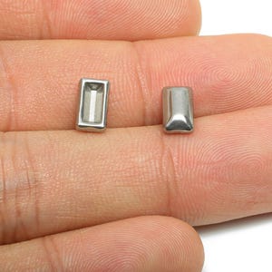 Puede incluir: Dos pequeños pendientes de botón rectangulares de color plateado. Un pendiente tiene una incrustación rectangular blanca. Los pendientes se muestran sobre la piel de una persona.