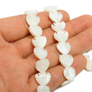 May include: Strands of white, heart-shaped beads with a carved design. The beads are held in a hand against a white background. The beads are made of a pearlescent material.