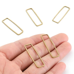 May include: A collection of gold-coloured rectangular metal jewellery findings. The open rectangles are held in a hand against a white background. These findings are likely used for jewellery making, such as earrings or pendants.