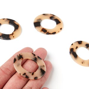 Peut inclure: Quatre perles ovales en résine avec un motif écaille de tortue. Les perles sont de couleur beige clair avec des taches marron foncé et noires. Une perle est tenue dans une main. Idéal pour la création de bijoux.
