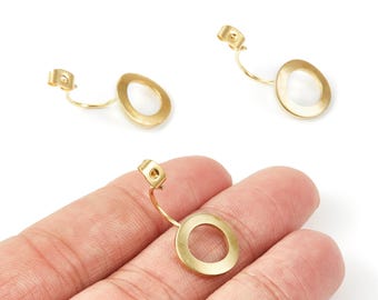 May include: Gold-toned earrings featuring an open oval shape. Each earring has a post and a curved connecting element. One earring is held in a hand, with two others positioned on a white backdrop. The earrings are likely made of metal.