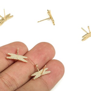 Op de afbeelding: Goudkleurige oorstekers in de vorm van een libel. De oorbellen hebben een gedetailleerd libellenontwerp met getextureerde vleugels en een pin voor doorboorde oren. Er zijn meerdere oorbellen zichtbaar.