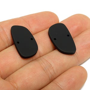 Könnte beinhalten: Zwei schwarze, oval geformte Komponenten mit flacher Oberfläche und einem kleinen Loch oben. Die Komponenten werden in einer Hand gehalten. Die Komponenten scheinen aus einem matten Material zu bestehen.