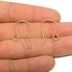 Può includere: Sono mostrati due ganci per orecchini color oro. Ogni gancio ha una parte superiore curva con una piccola sfera e un fondo dritto e appuntito. I ganci sono tenuti in una mano su sfondo bianco, mostrando componenti per gioielli.