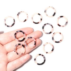 May include: A collection of ten round, open-center beads with a tortoiseshell pattern. The beads are a mix of cream, brown, and black, and are displayed on a white background. These beads are suitable for jewelry making.