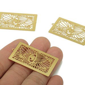 May include: Three gold-colored rectangular metal charms with an openwork floral design. One charm is held in a hand, while the other two are on a white background. Each charm has a small loop at the top for attaching to jewelry.