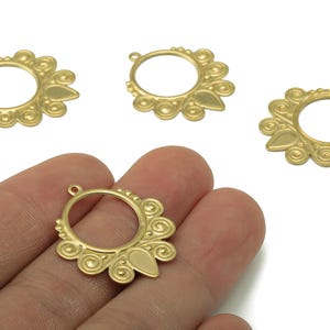 May include: Gold-colored filigree earring components with a circular design, featuring decorative swirls and a teardrop-shaped element. One component is held in a hand, with three others displayed nearby. These components are ideal for jewelry making.