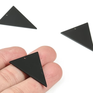Pode incluir: Três pingentes pretos em forma de triângulo com um pequeno orifício para enfiar. Um pingente é segurado em uma mão. Os pingentes são de cor preta fosca e estão sobre um fundo branco.