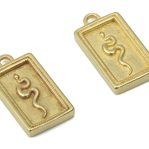 Puede incluir: Dos dijes de metal dorado en forma de rectángulo con un diseño de serpiente en relieve en el interior. Los dijes tienen un bucle en la parte superior para sujetarlos a un collar o pulsera.