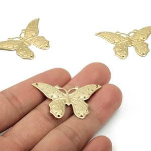 Puede incluir: Tres dijes de mariposa dorados con patrones de alas detallados. Uno se sostiene en una mano, mostrando el intrincado diseño. Las mariposas tienen pequeños agujeros para sujetar a joyas o manualidades.