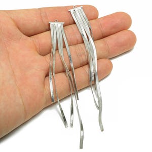 Puede incluir: Pendientes plateados con un diseño de flecos en cascada. Cada pendiente presenta múltiples hebras de tiras metálicas planas y rectangulares, creando una apariencia fluida y texturizada. Los pendientes se sostienen en una mano sobre un fondo blanco.