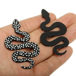 May include: Two snake-shaped accessories. One is black with white polka dots, the other is solid black. The accessories are likely earrings or pendants. The snake designs are stylized and have a smooth finish.