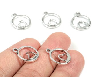 May include: Silver-colored heart charms. Each charm features a heart shape within a circular frame, with a small loop at the top for attachment. The charms are shown in a hand against a white background. These charms are suitable for jewelry making.