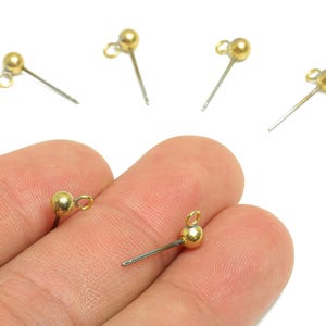 Könnte beinhalten: Nahaufnahme von mehreren goldfarbenen Ohrringsteckern mit kleinen, runden Perlen und angebrachten Schlaufen. Die Stifte sind silberfarben. Ein Ohrringstift wird zwischen den Fingern gehalten. Der Hintergrund ist weiß.