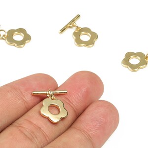 May include: Gold-coloured flower-shaped toggle clasps. The clasps have a bar and a flower-shaped loop. The image shows the clasps on a white background. The clasps are small and delicate, suitable for jewellery making.