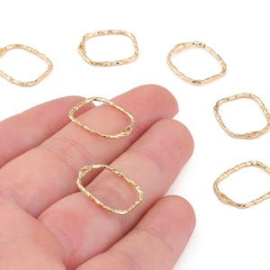 May include: Gold-colored, open-frame rectangular jewelry findings. The findings have a textured, organic appearance. Several pieces are scattered on a white surface, with a few held in a hand. These findings are suitable for jewelry making.