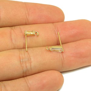 May include: Two gold-colored stud earrings with rectangular, clear gemstones. The earrings are held in a hand, showcasing their design and the post and hook details. The earrings are small and delicate.