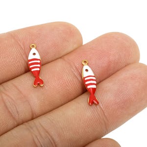 Può includere: Due piccoli ciondoli a forma di pesce. Ogni ciondolo è rosso e bianco con accenti dorati. I pesci hanno una coda rossa e un anello dorato nella parte superiore per il fissaggio ai gioielli. I ciondoli sono tenuti in una mano.