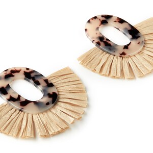 May include: A pair of oval-shaped earrings with a brown and white tortoiseshell pattern. The earrings have a fringe of beige raffia around the bottom.