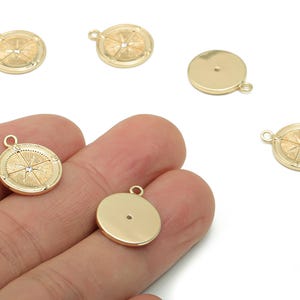 Op de afbeelding: Goudkleurige ronde bedelhangers met een kompasontwerp. De bedels hebben een kleine lus aan de bovenkant om aan sieraden te bevestigen. Eén bedel wordt in een hand gehouden. Het kompasontwerp heeft lijnen die uitstralen vanuit een centraal punt, met een klein kristallen accent.