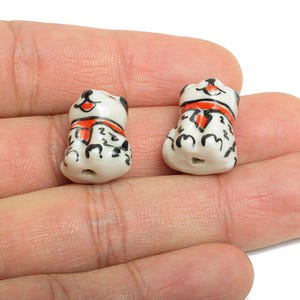 May include: Two small, white ceramic dog figurines with black spots and accents. Each dog wears a red scarf and has a smiling face. The figurines are approximately 2 cm tall and are designed with a hole for stringing.
