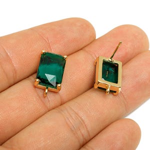 May include: A pair of emerald green rectangular stud earrings with gold-colored frames. The earrings have a faceted gemstone design and a post backing. The earrings are displayed on a light background.