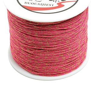 Könnte beinhalten: Eine Spule mit pinkfarbenem und goldenem Garn mit dem Text "DUOCAIJIEYI" auf dem Etikett.