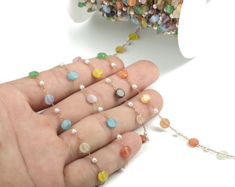Puede incluir: Una cadena de cuentas de piedras preciosas de colores con pequeñas perlas blancas y una cadena dorada. Las cuentas son en tonos de azul, verde, amarillo, naranja y rosa. La cadena se sostiene en una mano sobre un fondo blanco.