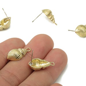 May include: Gold-colored seashell stud earrings. The earrings are in the shape of a conch shell with detailed ridges. One pair is held in a hand, while others are scattered on a white background. The earrings have a post and a small loop.