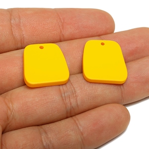 May include: Two bright yellow, rectangular pendants with rounded edges and a small hole at the top. The pendants are displayed on a hand, showcasing their color and shape. These pendants are suitable for jewelry making.