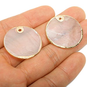 May include: Two round, pale pink pendants with a gold-coloured rim and a small hole at the top. The pendants have a pearlescent effect and are held in a hand.