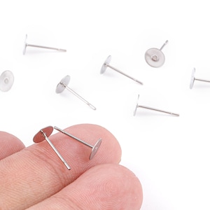 May include: A selection of silver earring posts, each with a flat, circular pad. The posts are straight with a small, round pad for attaching earrings. Several posts are scattered on a white background, with two held by a person's fingers.
