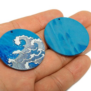 May include: Two round, blue pendants with a hole at the top. One pendant features a white and blue wave design. The other pendant is solid blue. These pendants are suitable for jewellery making.