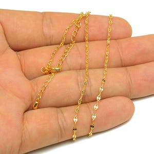 May include: A delicate gold-colored necklace with a chain of small, flat, oval-shaped links. The necklace is held in a hand, showcasing its fine details and adjustable length. The jewelry is designed for a minimalist aesthetic.