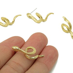 Könnte beinhalten: Goldfarbene Ohrstecker in Schlangenform. Die Ohrringe sind mit strukturierten Schuppen und einem Steckerverschluss versehen. Ein Ohrring wird in einer Hand gehalten, drei weitere sind auf einer weißen Oberfläche verstreut.