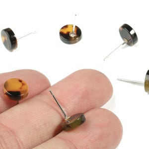 Könnte beinhalten: Ohrstecker mit Schildpattmuster. Die Ohrringe haben ein flaches, kreisförmiges Design mit einem dunkelbraunen und bernsteinfarbenen Farbschema. Die Stifte sind silberfarben. Mehrere Ohrringe sind auf weißem Hintergrund verstreut.