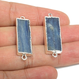 Puede incluir: Dos colgantes rectangulares de piedra preciosa de cianita azul con borde de metal plateado. Cada colgante tiene un lazo en la parte superior e inferior para sujetarlo a una cadena u otro componente de joyería. La cianita tiene un borde natural y rugoso.